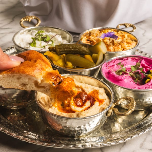 A hand dips flatbread into hummus, surrounded by four other Mediterranean dips and pickles on a silver tray.
